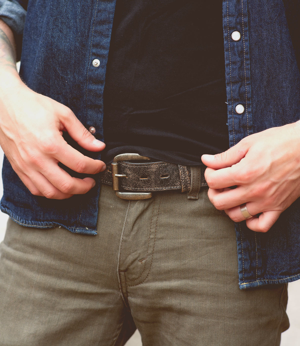 A person dressed in a denim shirt and dark pants adjusts their Bed Stu Meander made of genuine distressed leather with a removable antique metal buckle using both hands.