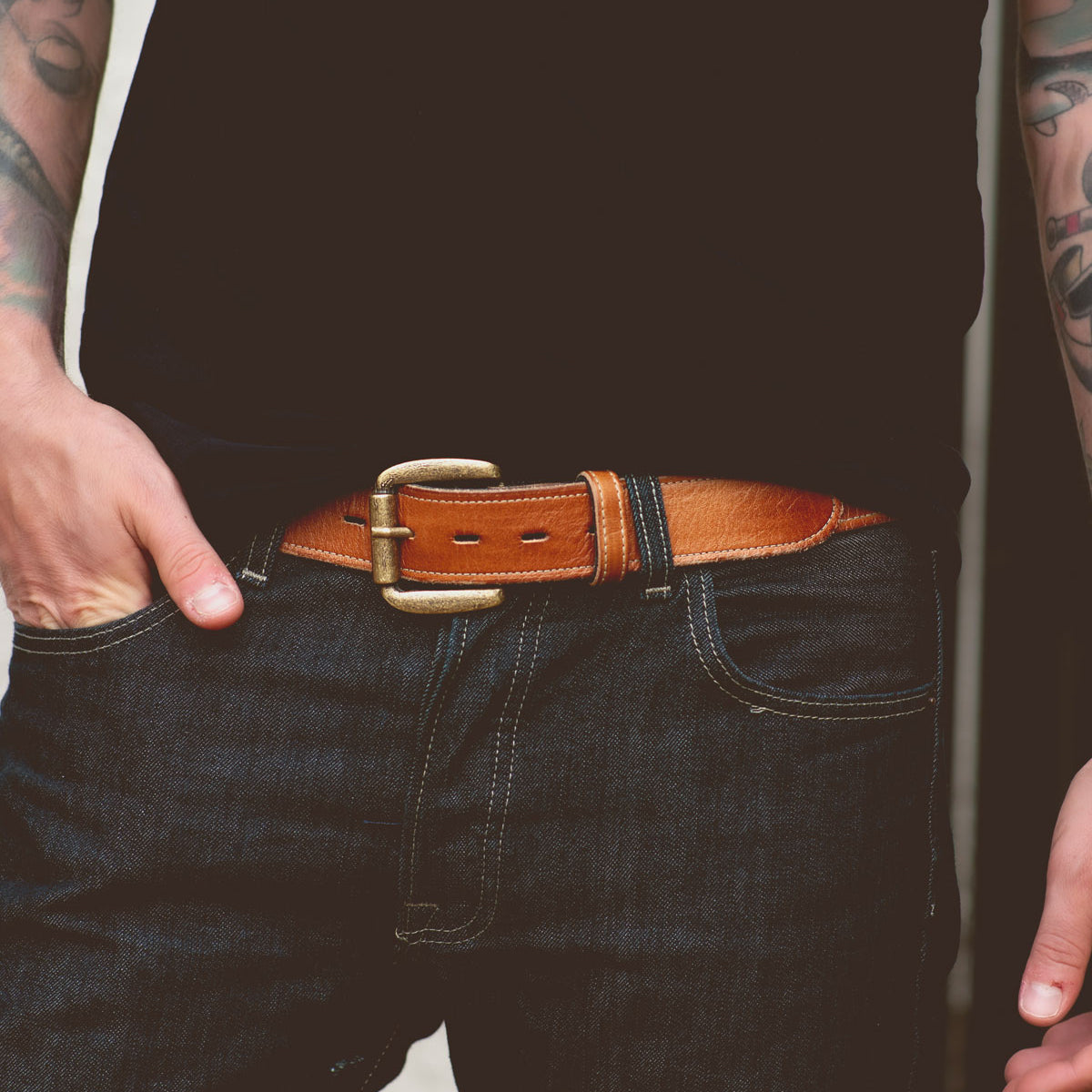 A person wearing a black shirt and dark jeans with a Meander belt by Bed Stu featuring a removable buckle and tattoos on their arms.