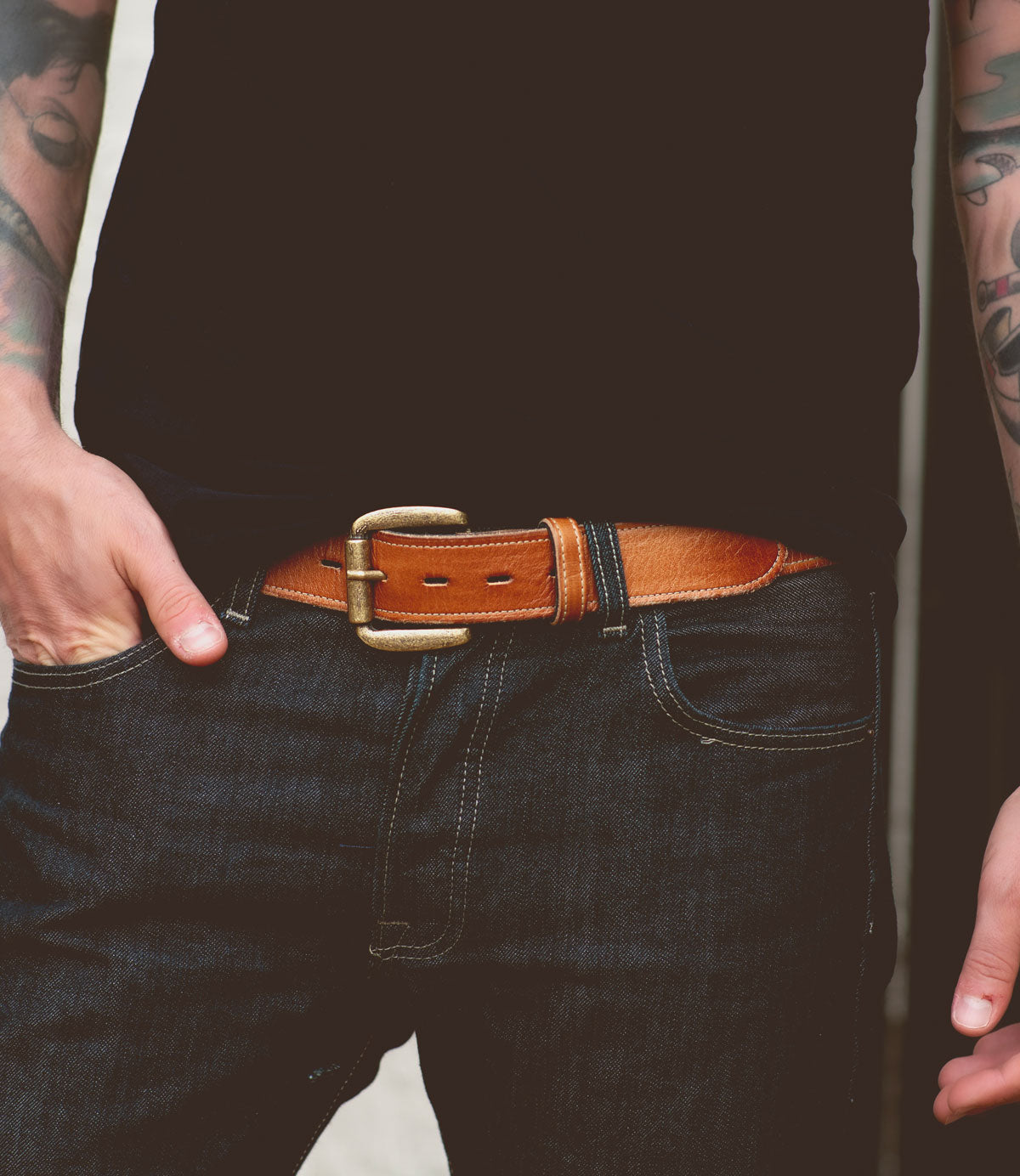A person wearing a black shirt and dark jeans with a Meander belt by Bed Stu featuring a removable buckle and tattoos on their arms.