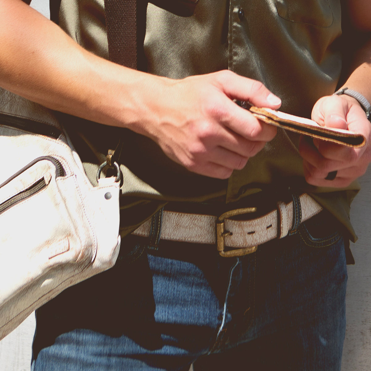 A person in a green shirt and blue jeans is holding a distressed leather wallet. They are also carrying a Meander white shoulder bag by Bed Stu.