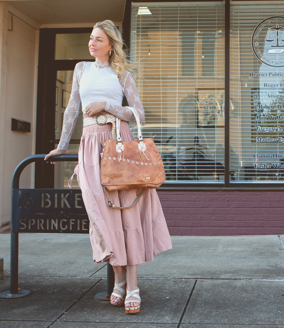 A woman in a white top and pink skirt standing on a sidewalk and leaning on a metal railing with a Bed Stu brown bag over her shoulder, wearing comfortable sandals.