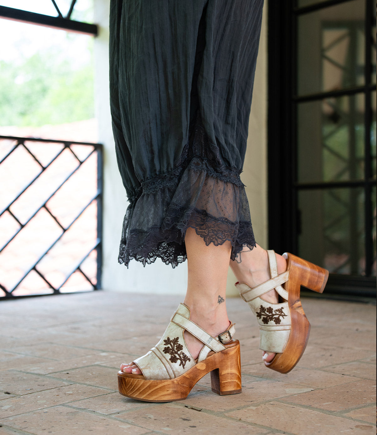A person wearing Bed Stu’s Melodramatic black lace-trimmed pants and beige platform sandals with a chunky leather heel and floral embroidery, standing on a tiled floor.