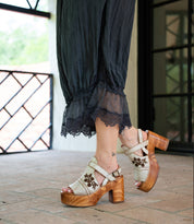 A person wearing Bed Stu’s Melodramatic black lace-trimmed pants and beige platform sandals with a chunky leather heel and floral embroidery, standing on a tiled floor.