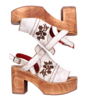 The Melodramatic by Bed Stu is a pair of vintage-inspired platform sandals with chunky leather heels, wooden soles, beige straps, and dark floral embroidery on the sides.