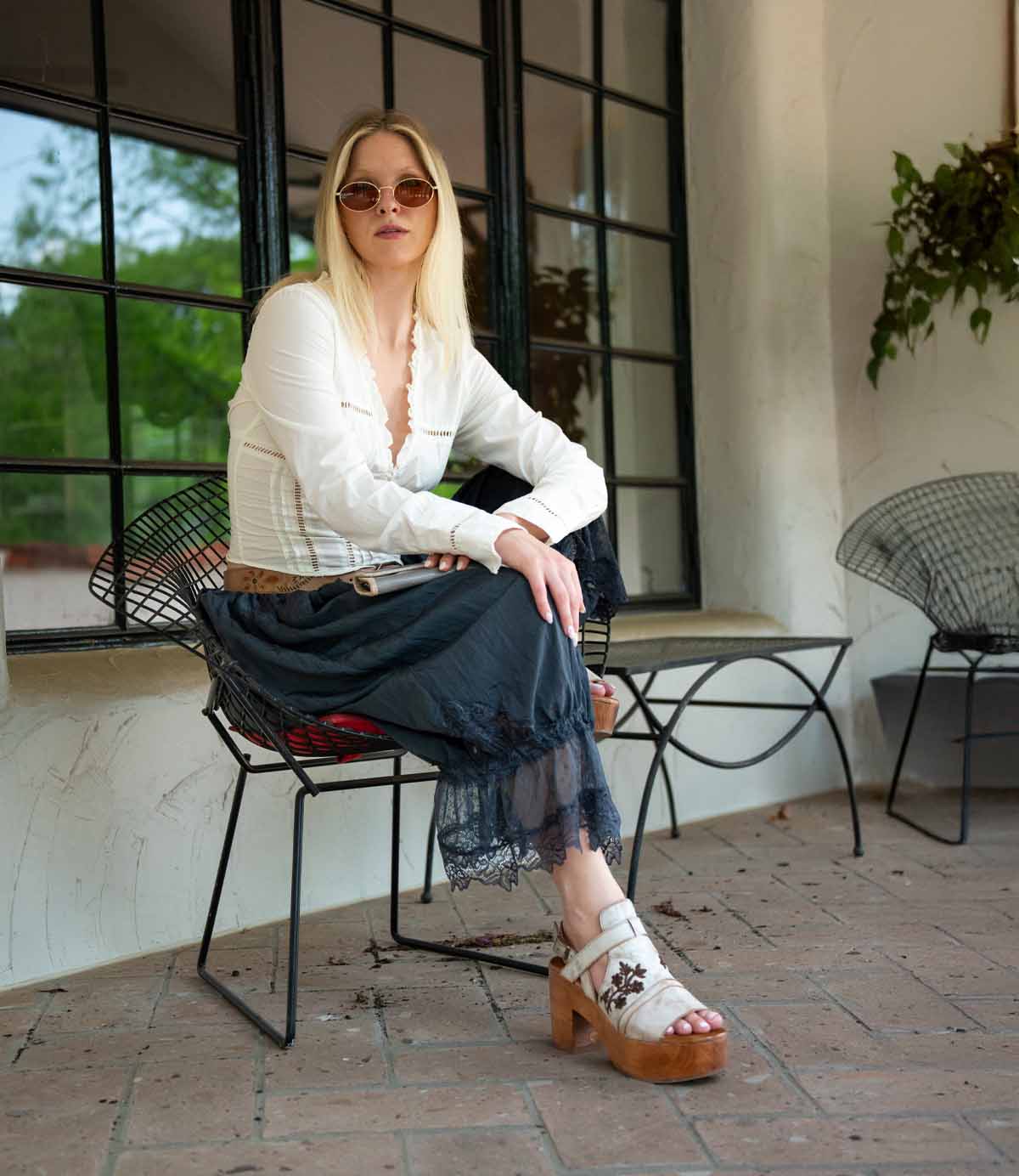 A woman with long blonde hair in sunglasses sits outdoors on a metal chair, wearing a white blouse, dark skirt, and Bed Stu’s Melodramatic platform sandals with embroidered leather.