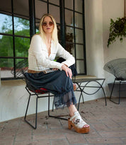 A woman with long blonde hair in sunglasses sits outdoors on a metal chair, wearing a white blouse, dark skirt, and Bed Stu’s Melodramatic platform sandals with embroidered leather.