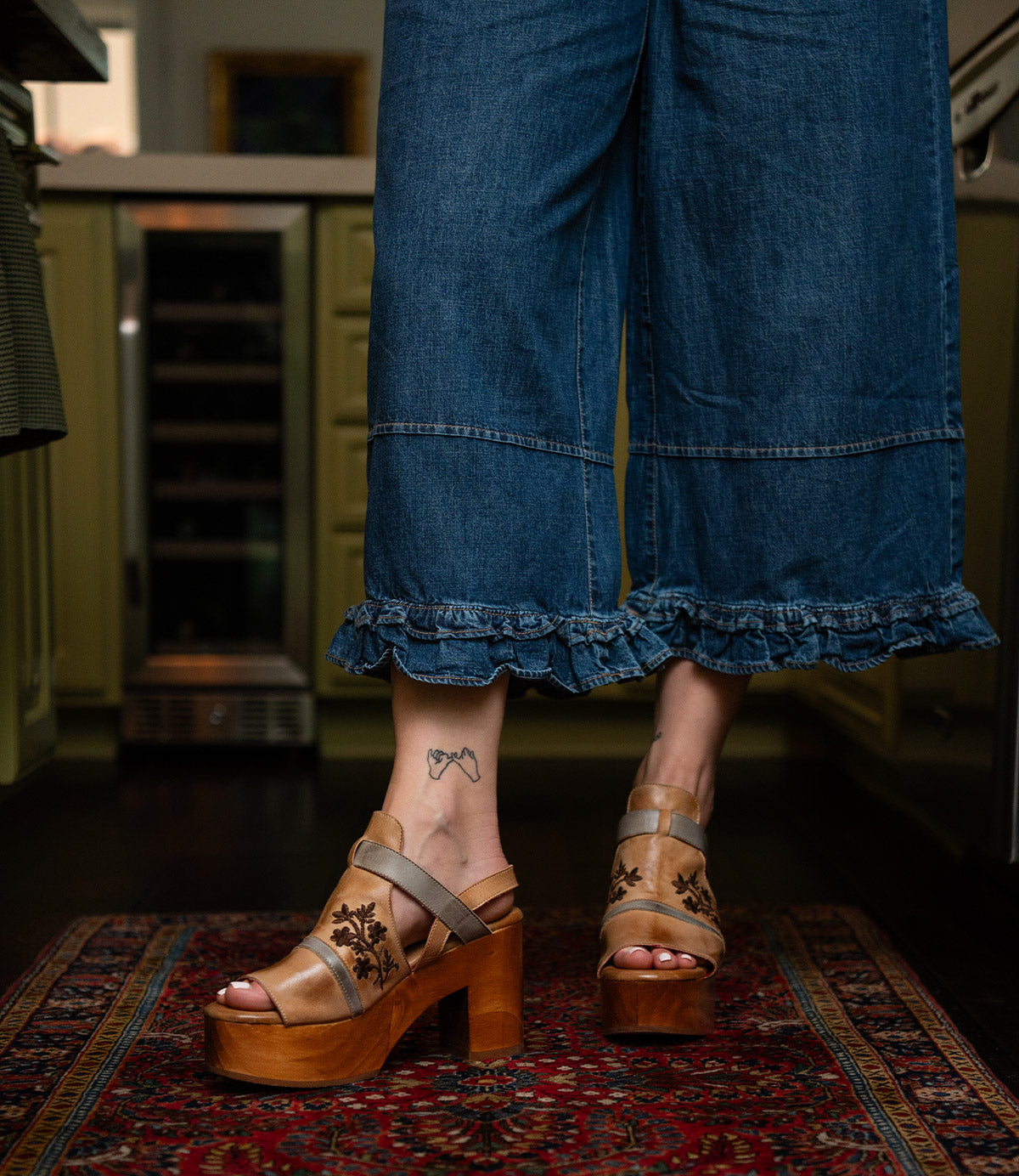 A person wears wide-leg denim pants with ruffled hems and Bed Stu’s Melodramatic tan platform sandals featuring a chunky leather heel, standing on a patterned rug. A butterfly tattoo is visible on their ankle.