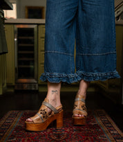 A person wears wide-leg denim pants with ruffled hems and Bed Stu’s Melodramatic tan platform sandals featuring a chunky leather heel, standing on a patterned rug. A butterfly tattoo is visible on their ankle.