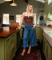 A woman stands in a kitchen wearing a brown corset top, wide-leg jeans with ruffled hems, and Bed Stu's Melodramatic platform sandals; a brown bag sits on the counter beside her.