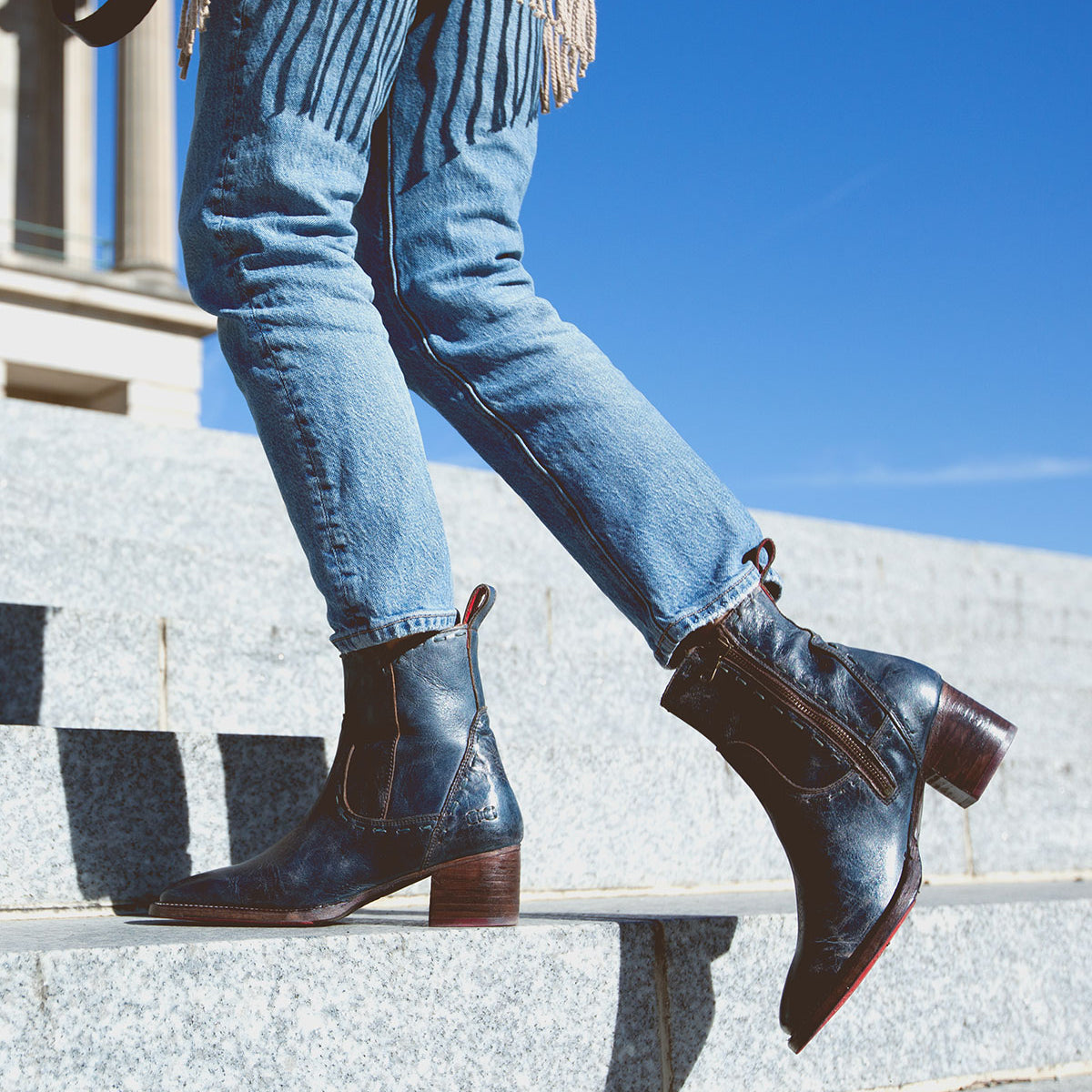 A person wearing blue Bed Stu Merryli leather boots with cushioned insoles is walking up stone steps outdoors under a clear blue sky.