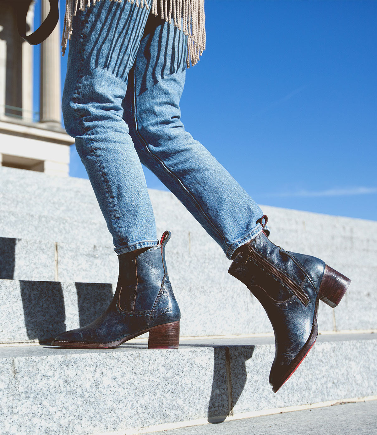 A person wearing blue Bed Stu Merryli leather boots with cushioned insoles is walking up stone steps outdoors under a clear blue sky.