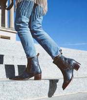 A person wearing blue Bed Stu Merryli leather boots with cushioned insoles is walking up stone steps outdoors under a clear blue sky.