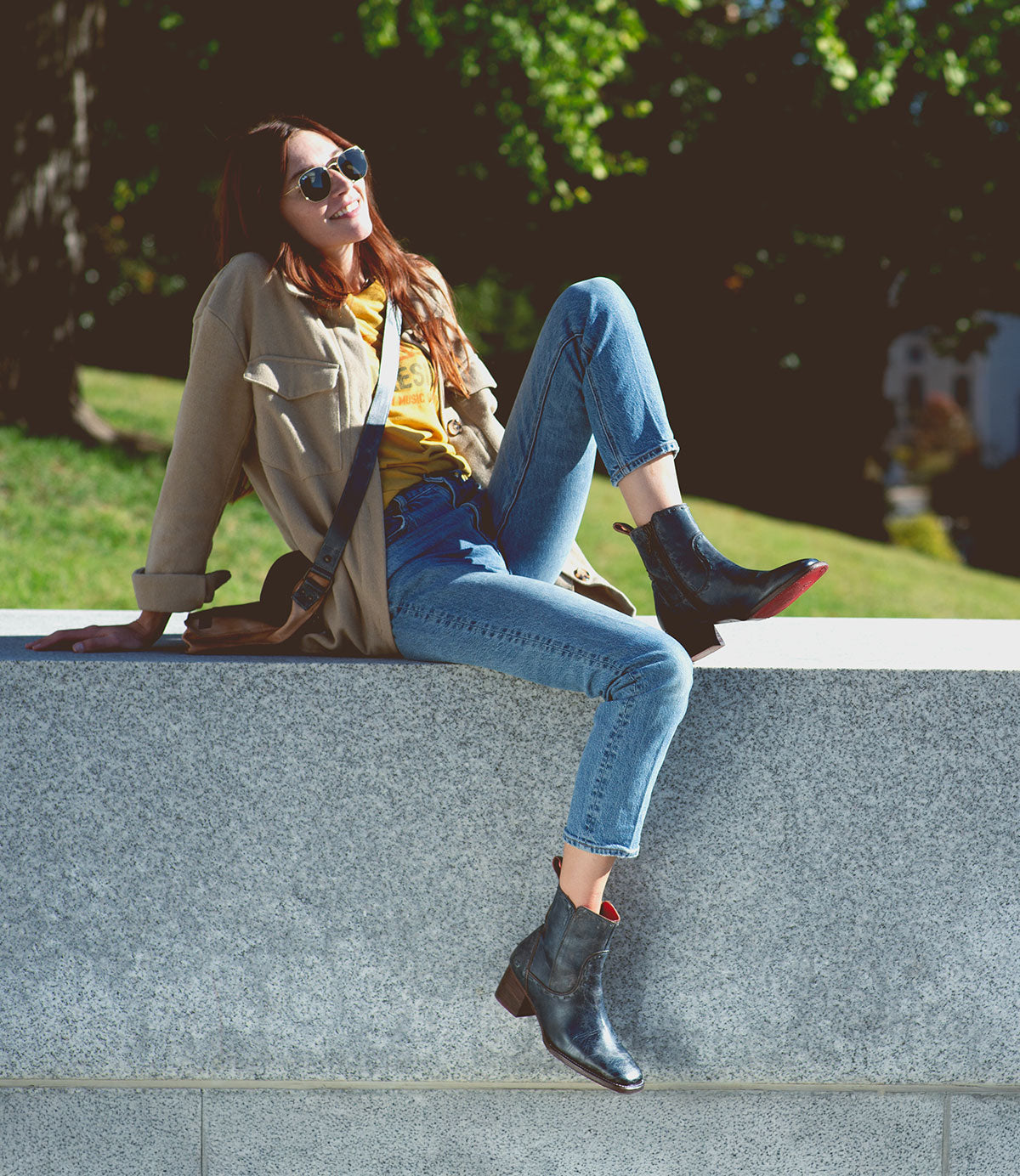 A person wearing stylish Merryli sunglasses by Bed Stu, a beige jacket, and jeans with cushioned insoles relaxes on a stone wall in a sunny outdoor setting.