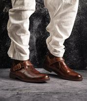 A person wearing the Bed|Stü Michelangelo brown leather dress shoes with a buckle accent and off-white pants stands on a gray textured floor with a dark background.