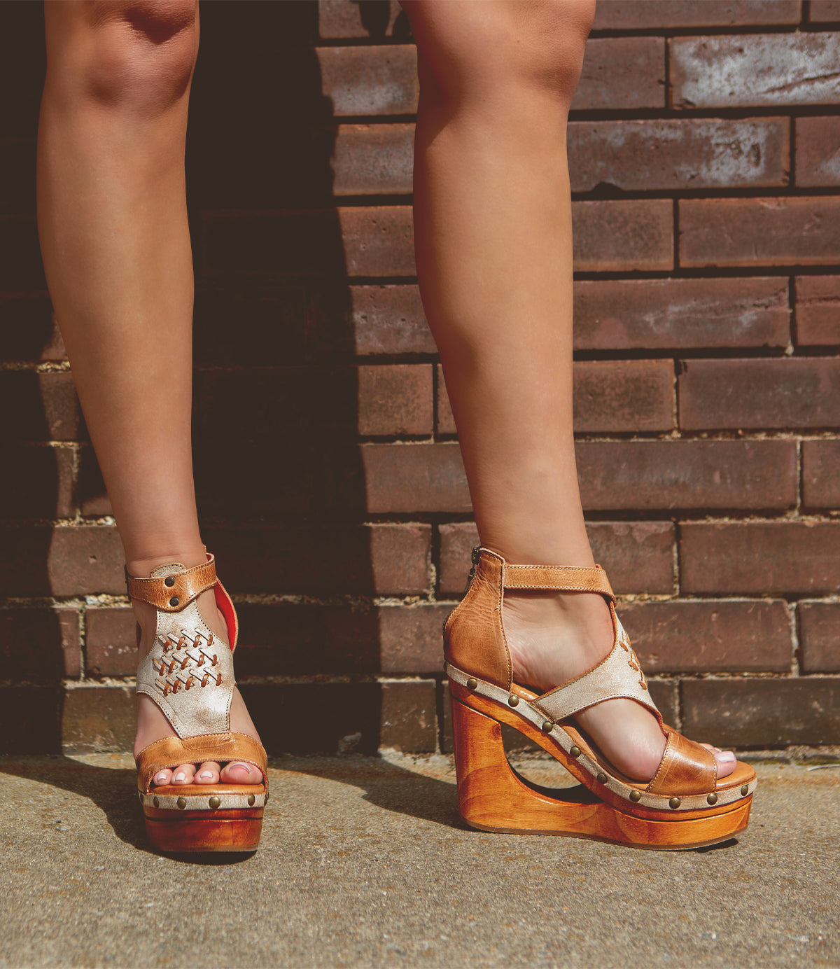 A person standing against a brick wall wearing brown Bed Stu Millennial t-strap front platform sandals.