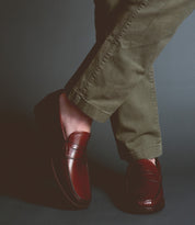A person wearing brown Bed Stu Moat loafers and green pants, with legs crossed, stands against a dark background, exuding timeless sophistication.