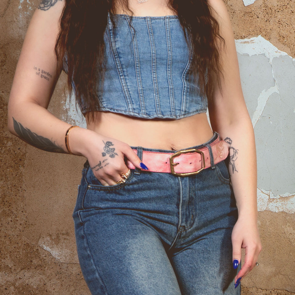 Wearing a Bed Stu Mohawk outfit, someone stands against a textured wall in a denim ensemble with a sleeveless top, high-waisted jeans, and a distressed leather belt. Tattoos and dark, wavy hair enhance the rugged charm.