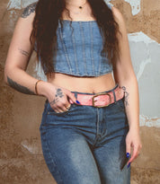 Wearing a Bed Stu Mohawk outfit, someone stands against a textured wall in a denim ensemble with a sleeveless top, high-waisted jeans, and a distressed leather belt. Tattoos and dark, wavy hair enhance the rugged charm.