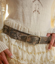 A person wearing a cream knit sweater and skirt styled with the Bed Stu Mohawk women’s floral belt, featuring a wide, worn leather strap and painted floral design.