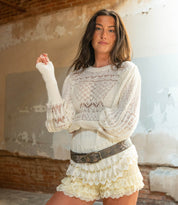 A woman stands indoors against a worn wall, wearing the Bed Stu Mohawk white knit sweater with puffy sleeves and cream lace shorts, styled with a chic women's floral belt.