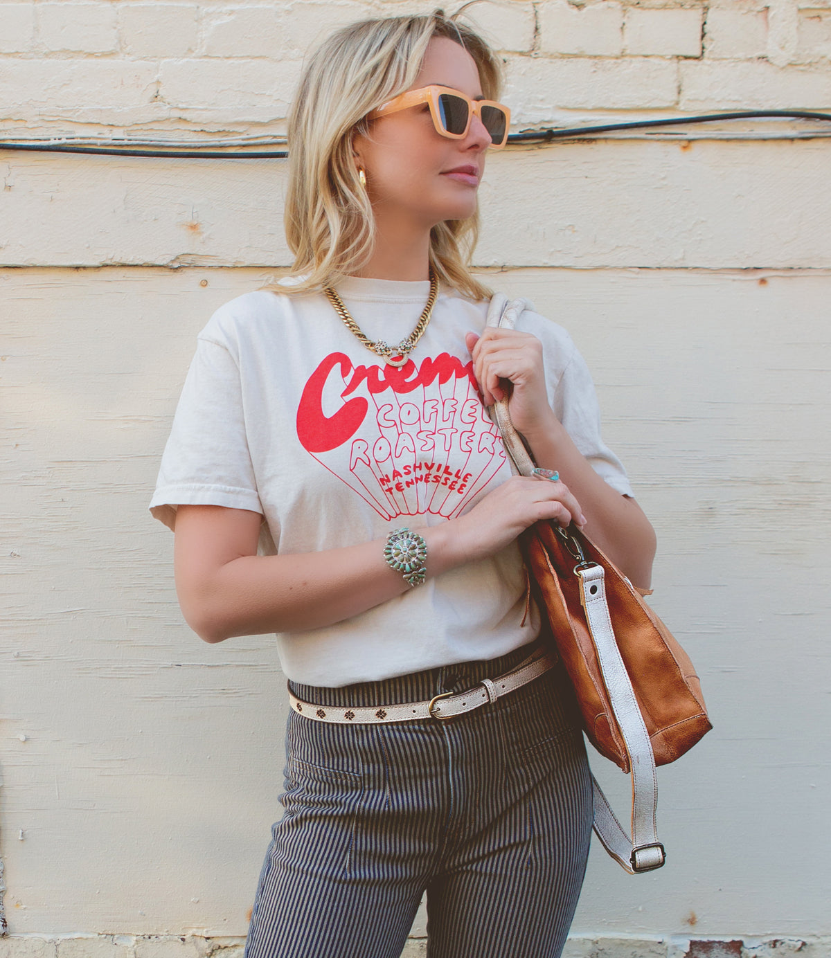 Woman in a casual outfit with a graphic t-shirt and striped trousers, accessorized with sunglasses, a watch, a Bed Stu Monae II premium leather belt with hand-stamped details, and a shoulder bag, standing against a