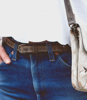 Close-up of a person wearing blue jeans, a white shirt, and a premium leather belt with hand-stamped details. They are holding the brown strap of a Bed Stu Monae II.