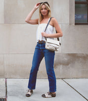 A woman stands on a sidewalk wearing a white top, blue jeans, and patterned shoes. She holds sunglasses on her head with one hand and has a refined accessory in the form of a Bed Stu Monae II crossbody bag on her shoulder.