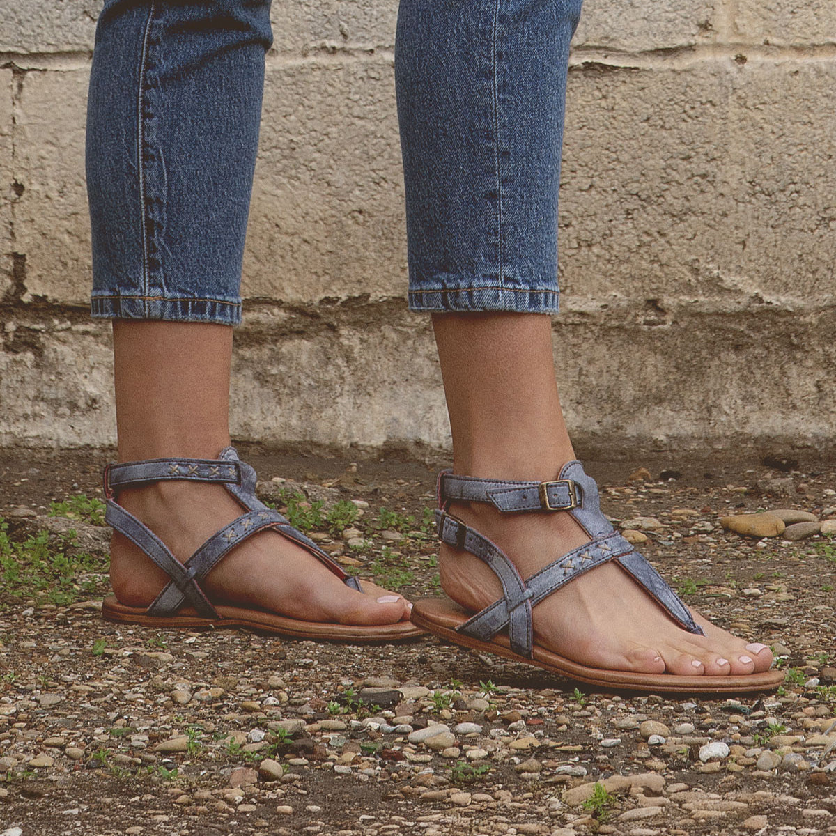 Someone wearing blue denim jeans and Bed Stu's Moon artisanal craftsmanship blue strappy sandals stands on gravel, with a concrete wall in the background.