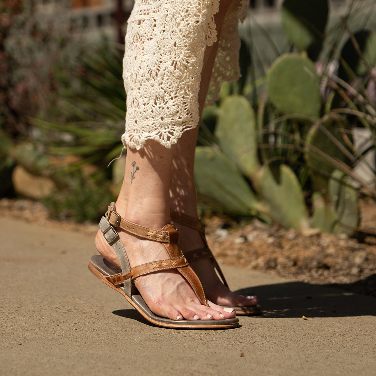 Wearing the cream Moon dress by Bed Stu and brown leather sandals with ankle buckles, a person walks on a sunlit concrete path lined with cacti.