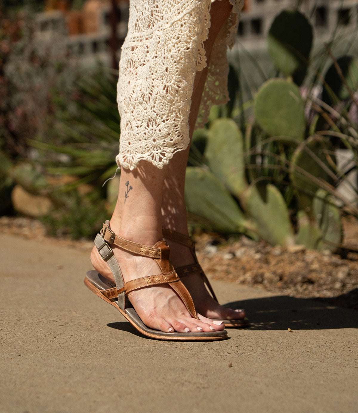 Wearing the cream Moon dress by Bed Stu and brown leather sandals with ankle buckles, a person walks on a sunlit concrete path lined with cacti.