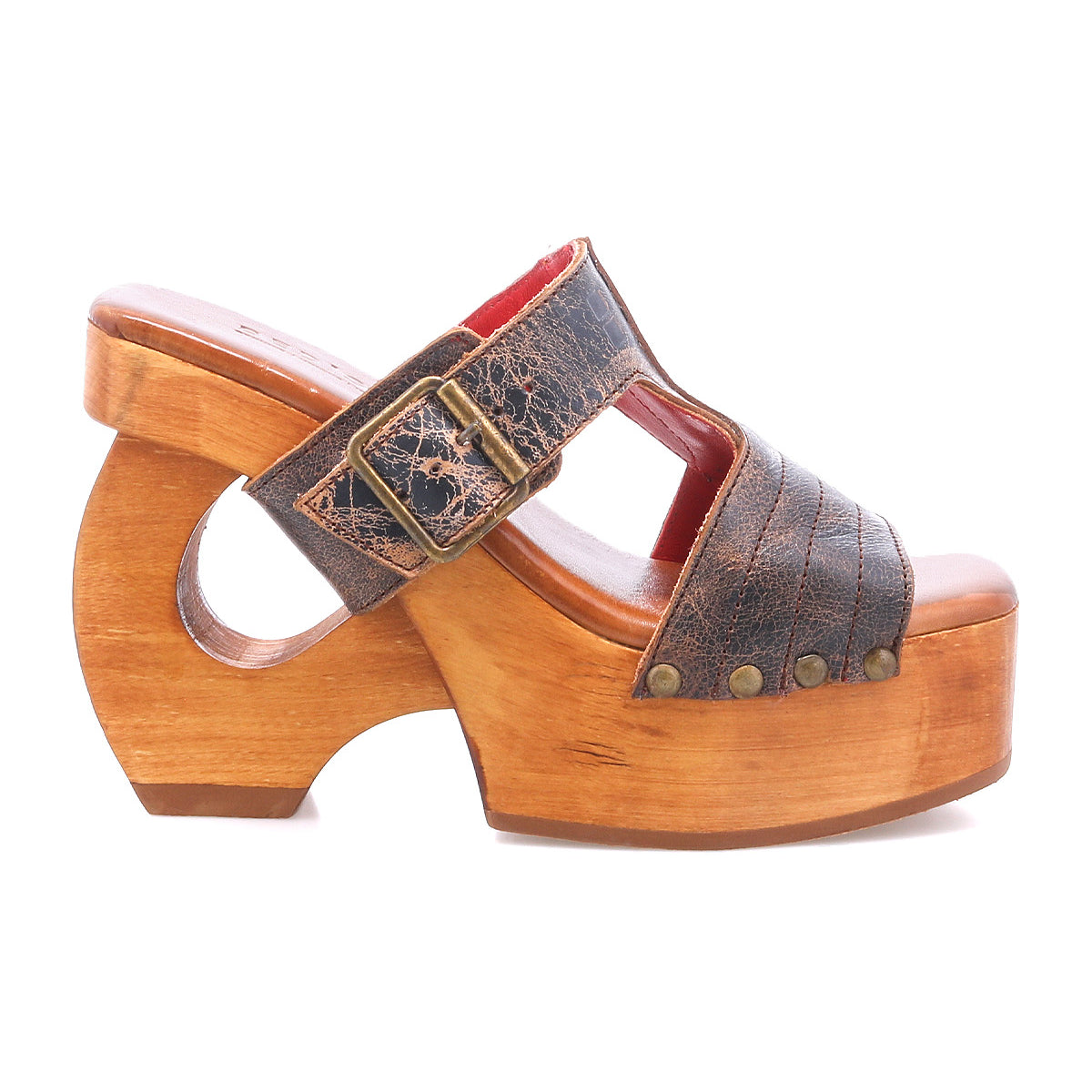 Mutable by Bed Stu is a wooden platform sandal with a curved wooden heel, brown leather strap, large decorative buckle, and decorative rivets.
