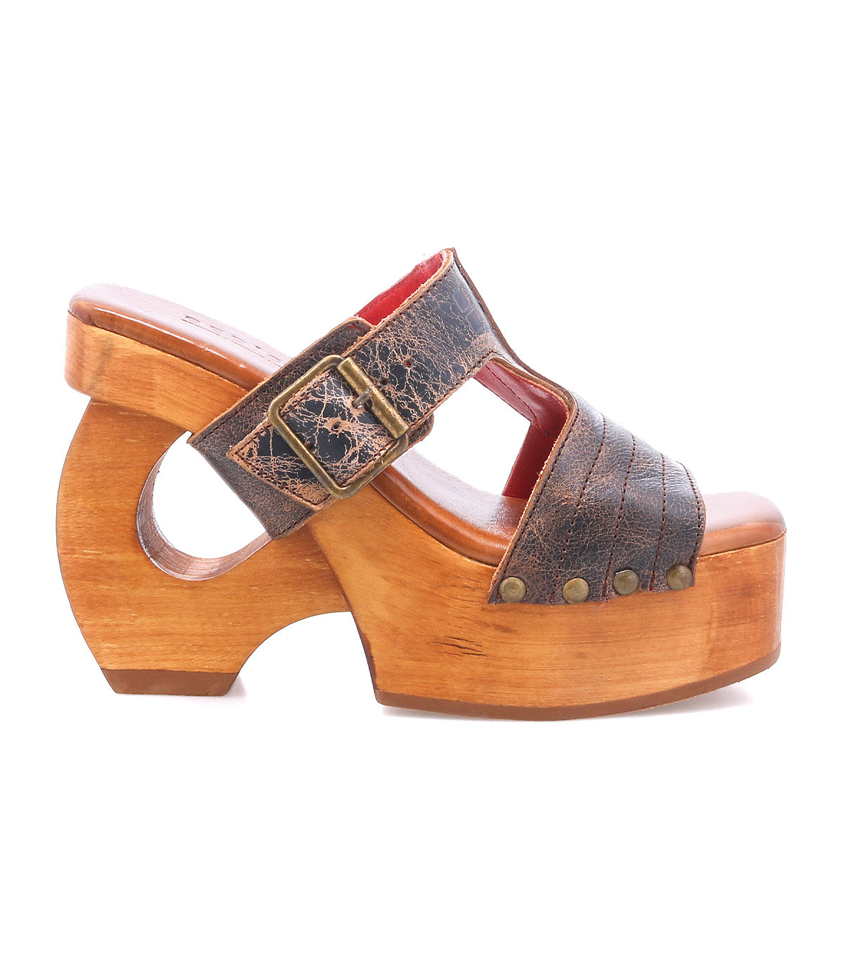 Mutable by Bed Stu is a wooden platform sandal with a curved wooden heel, brown leather strap, large decorative buckle, and decorative rivets.