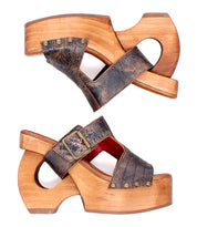 The Bed Stu Mutable sandals feature distressed leather straps, metal studs, and a decorative buckle on a vintage wooden platform, shown from the side against a white background.