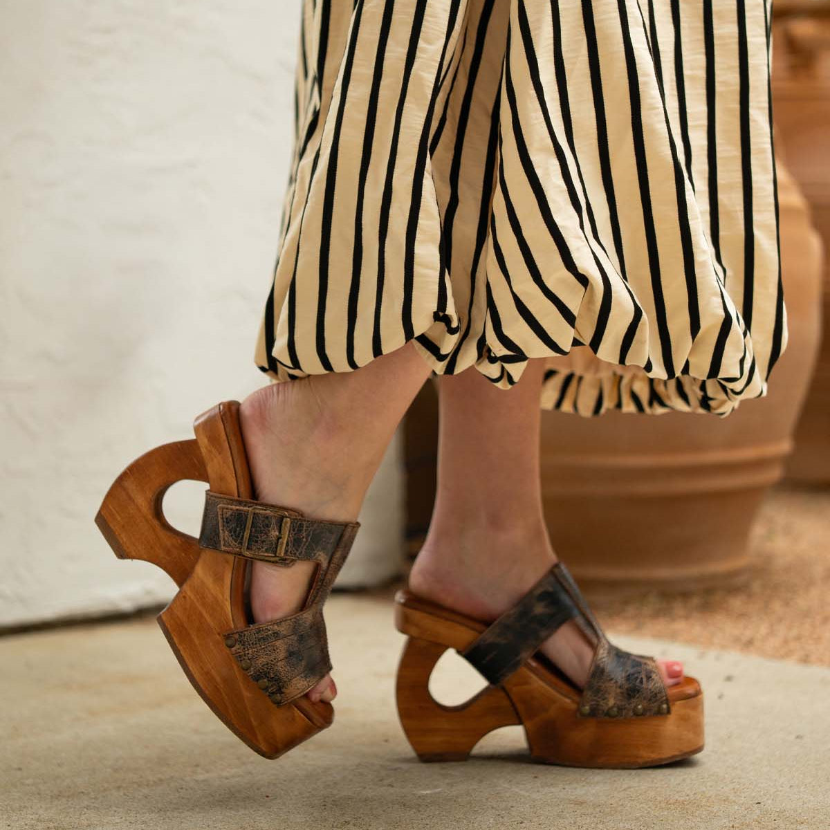 A person stands on a concrete floor near potted plants, wearing a long black and white striped skirt and Bed Stu's Mutable brown platform sandals with unique cut-out wooden heels that add a touch of vintage flair.