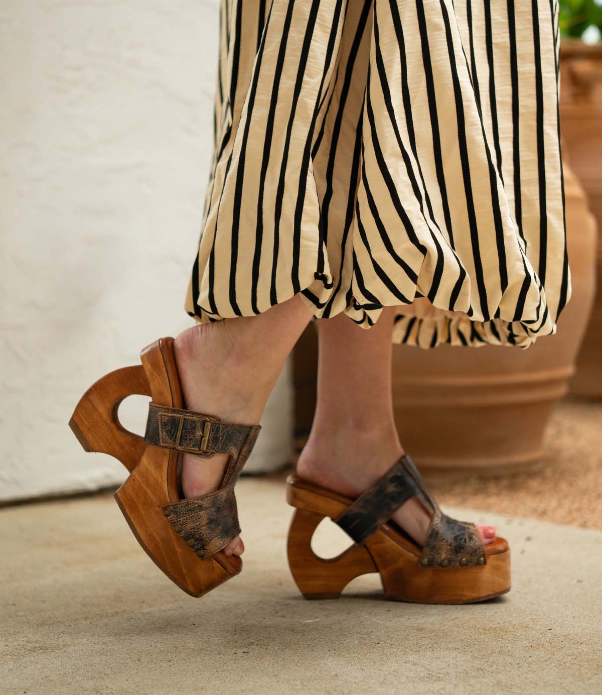 A person stands on a concrete floor near potted plants, wearing a long black and white striped skirt and Bed Stu's Mutable brown platform sandals with unique cut-out wooden heels that add a touch of vintage flair.