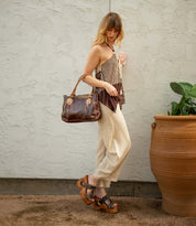 A woman stands outdoors next to a large clay pot and plant, holding the Bed Stu Mutable brown leather handbag with a decorative buckle, paired with a striped top, cream pants, and platform sandals.