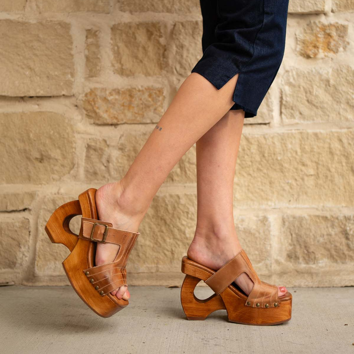 A person stands on concrete against a stone wall, wearing dark rolled-up pants and Bed Stu Mutable brown wooden platform sandals with a decorative buckle.