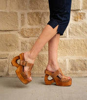 A person stands on concrete against a stone wall, wearing dark rolled-up pants and Bed Stu Mutable brown wooden platform sandals with a decorative buckle.