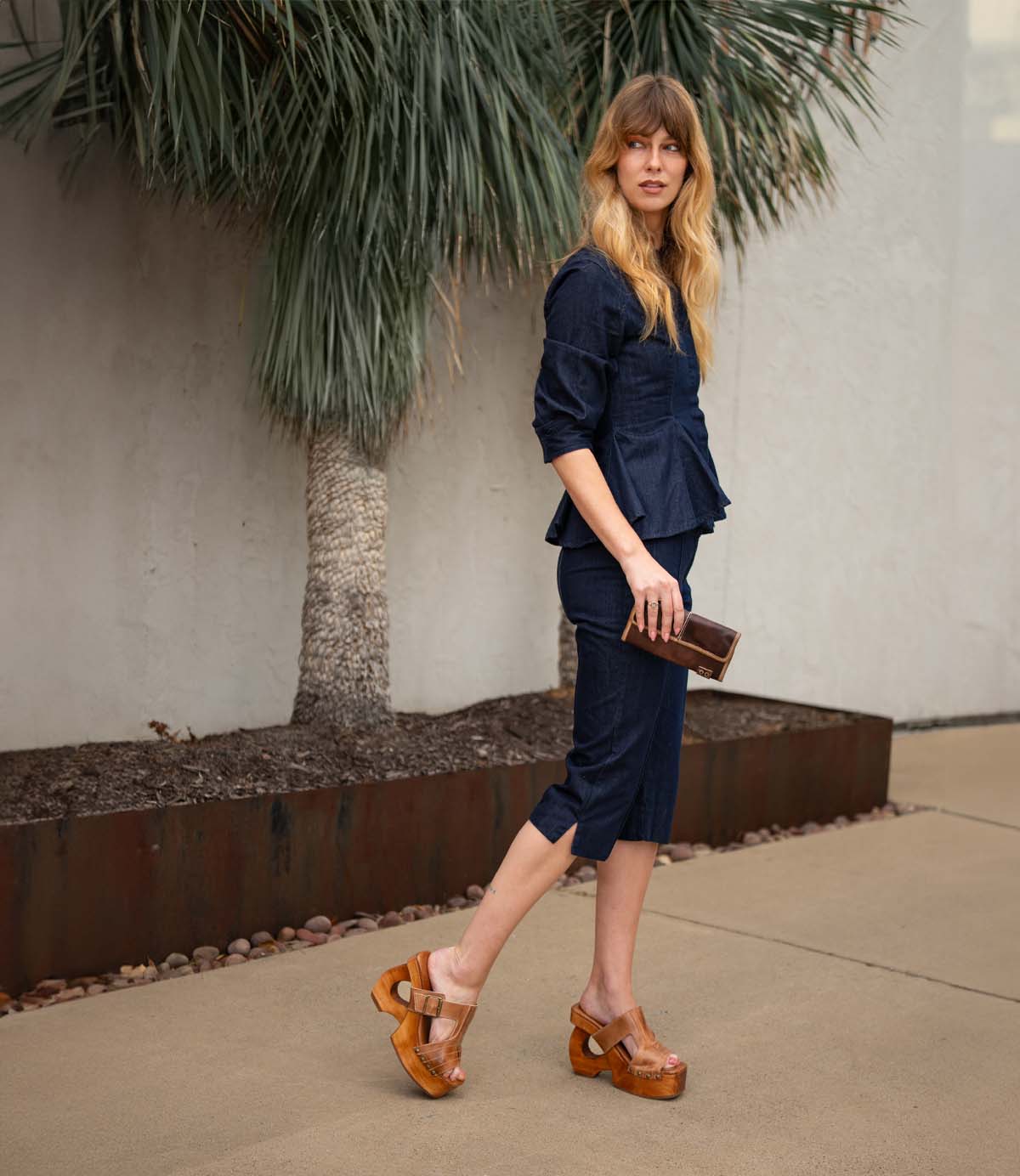 A woman wearing Bed Stu's Mutable dark blue set and wooden platform sandals with a decorative buckle, holding a brown clutch, walks by a palm tree.