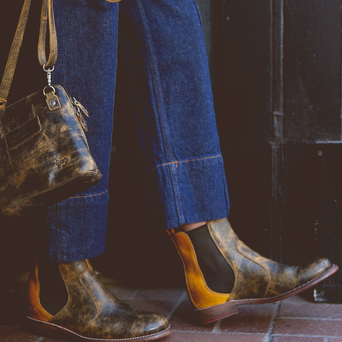 Person wearing brown Nandi Chelsea boots from Bed Stu, blue jeans, and carrying a worn leather handbag.