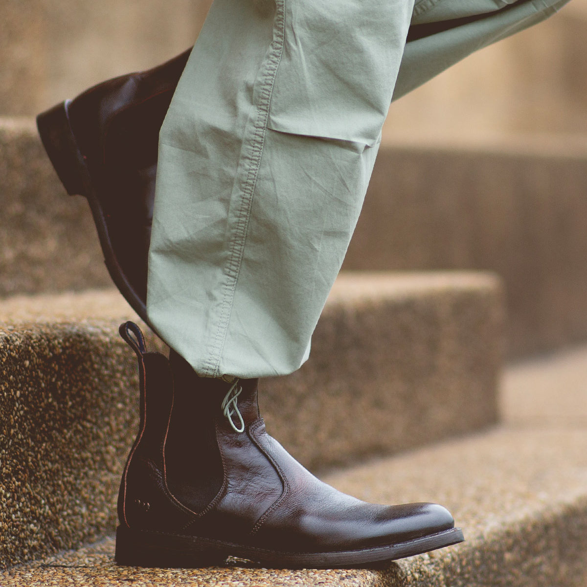 Close-up of a person's legs wearing light green pants and dark brown leather Bed Stu Nando ankle boots with a durable Vibram outsole, walking up stone steps.