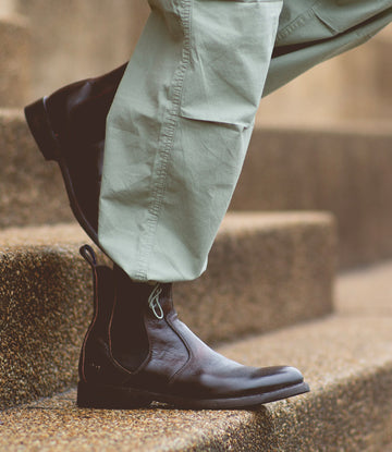 Close-up of a person's legs wearing light green pants and dark brown leather Bed Stu Nando ankle boots with a durable Vibram outsole, walking up stone steps.