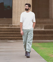 A man with a beard, wearing a white shirt and light green pants, walks on a paved path near stone steps and a grassy area. His look is completed with stylish Nando boots by Bed Stu featuring a Vibram® outsole, perfect for both comfort and durability.
