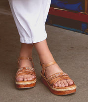 Close-up of a person wearing white pants and brown strappy sandals with thick soles, showcasing the latest in sandal fashion on the concrete surface. The chic design of Necessary by Bed Stu features stitched straps that add a touch of elegance to the look.