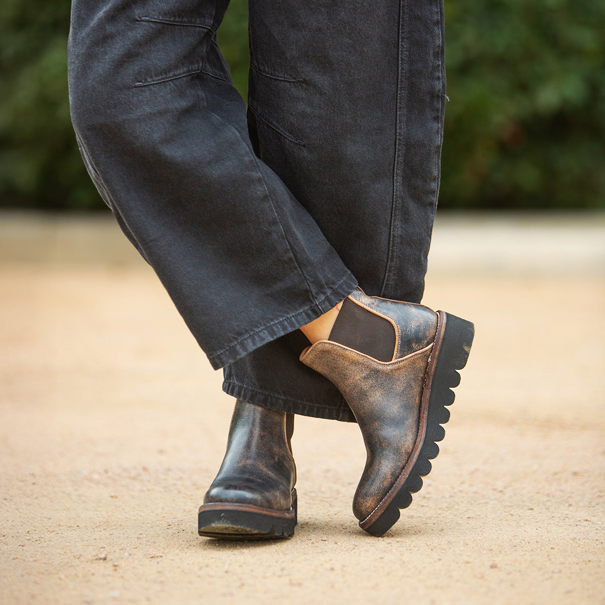 Wearing Bed Stu's Novel II distressed leather Chelsea boots with a lightweight sole and loose dark pants, someone stands on sandy ground with greenery behind them.