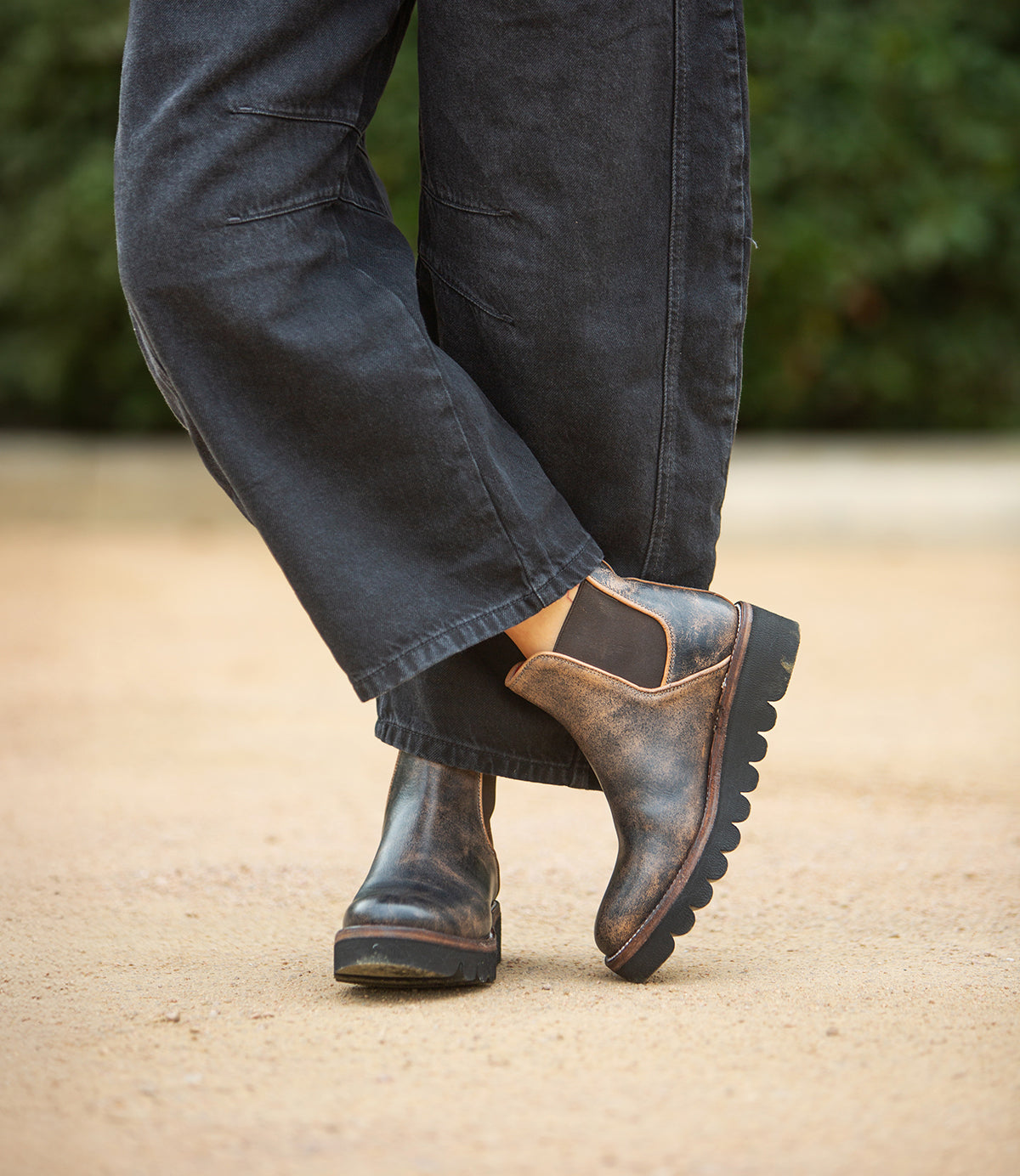 Wearing Bed Stu's Novel II distressed leather Chelsea boots with a lightweight sole and loose dark pants, someone stands on sandy ground with greenery behind them.