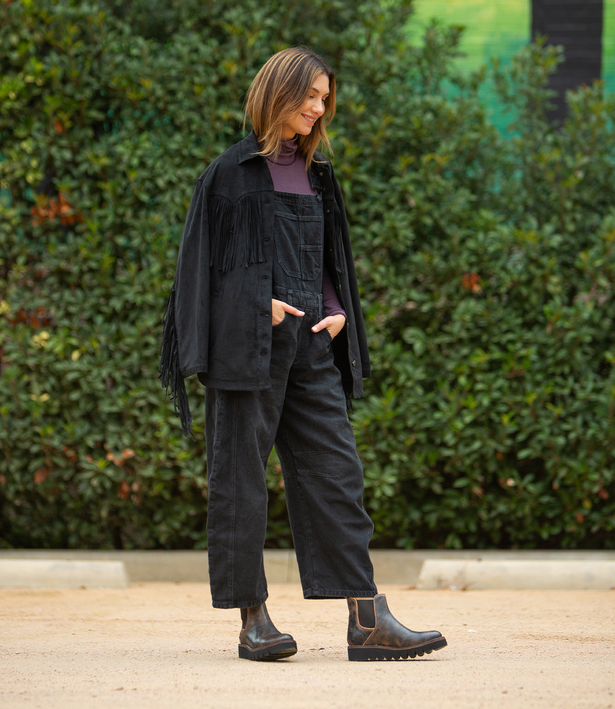 A woman stands outdoors on a dirt path amid green foliage, wearing black overalls, a fringed jacket, a purple turtleneck, and Bed Stu's Novel II Chelsea boots with a leather upper and lightweight sole.