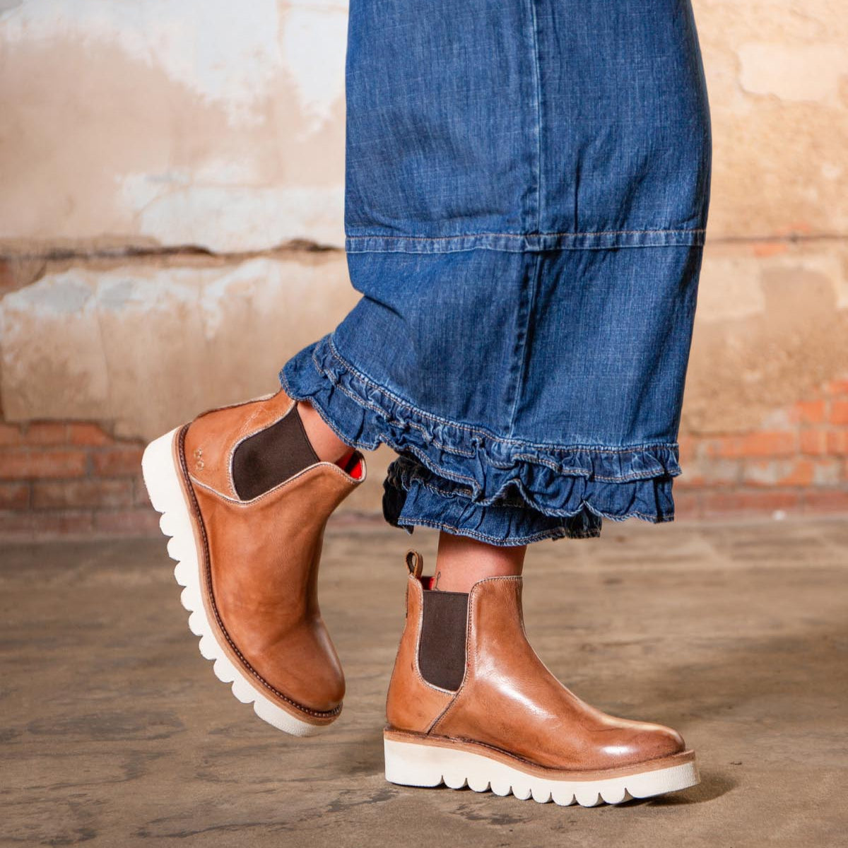 A person wearing cropped denim jeans with ruffled hems and Bed Stu Novel II tan leather Chelsea boots with a lightweight sole, standing on a concrete floor against a textured wall.