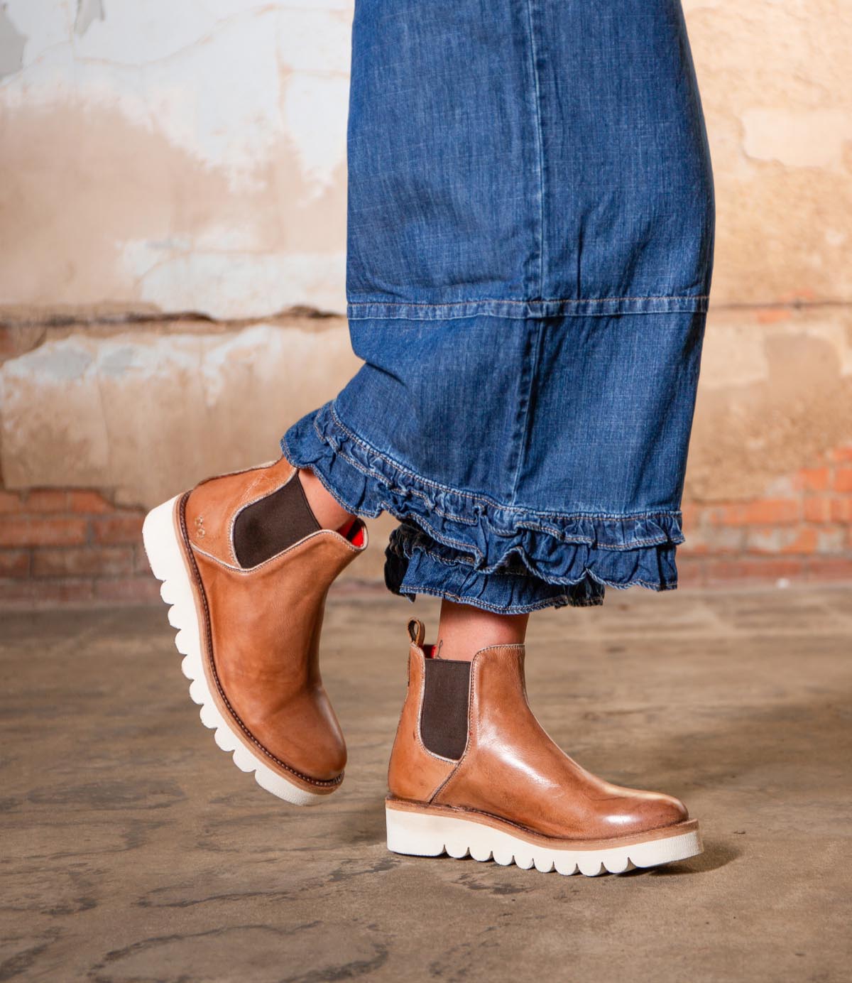 A person wearing cropped denim jeans with ruffled hems and Bed Stu Novel II tan leather Chelsea boots with a lightweight sole, standing on a concrete floor against a textured wall.