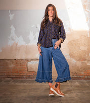 A woman stands indoors against a distressed wall, wearing a patterned blouse, wide-leg denim culottes, and Bed Stu’s Novel II brown shoes with a cushioned platform.